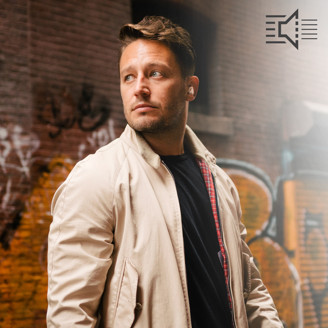 Man standing in front of a graffiti covered brick wall wearing a beige jacket and Klipsch T5 II True Wireless ANC Earphones mobile